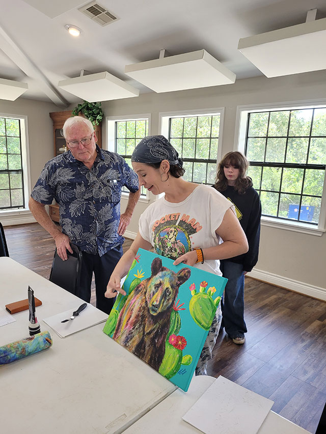 Photo of Virginia explaining her process on a colorful painting of a bear while 2 members look on.