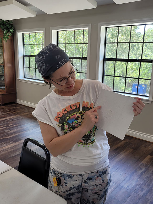 Photo of Virginia holding a canvas panel that has dried modeling paste on it, pointing out how it adds texture.