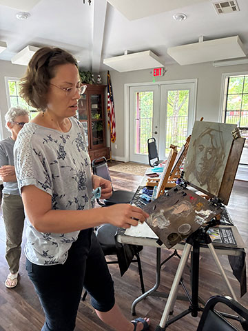 Photo of AnneMarie standing in front of an easel with a painting on it as one of the participants looks on.